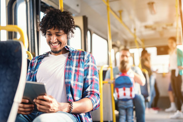Smiling African American guy sitting in public transportation and using tablet. On ears earphones. In background people sitting and standing.