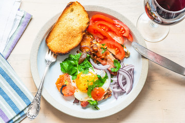 Fresh rustic breakfast - fried egg with bacon, onion and tomatoes on a plate and toast on a light wooden background, top view
