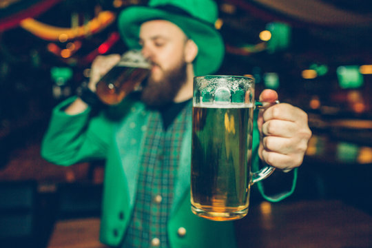 Young bearded man in green suit hold one mug of beer close to camera. He drink from another one. Guy stand in pub. He wear St. Patricks suit.