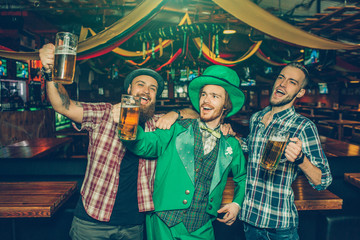 Happy three young men singing and waving with beer mugs in pub. They look to left. Guy in middle...