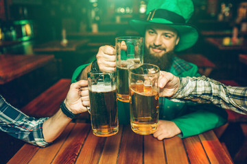 Picture of young man in green St. Patrick's suit sit at table with friends in pub. They hold mugs of beer together.
