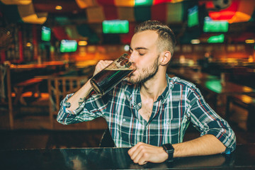 Young man sit lonely at bar counter in pub. He drink dark beer from mug.