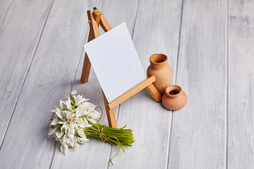 Bouquet of snowdrops and a small easel with a white paper and mini jars on agrey background. Greeting card with a space for text