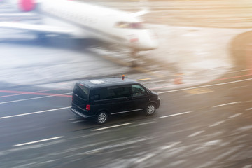 Black VIP service van running on airport taxiway with blurred private jet on background. Business class service at airport. Security intelligence agency hurrying at airfield