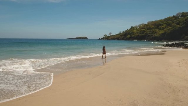 Young girl in a swimsuit walking on the beach. Aerial view ofBeautiful girl on sandy beach walking along the coast among the waves. Travel concept, Aerial footage, 4K video.