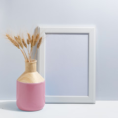 Mock up white frame and spikelets of wheat in pink vase on book shelf or desk. Minimalistic concept.
