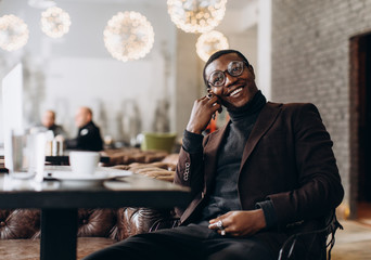 Portrait of happy african businessman using phone while working on laptop in a restaurant.