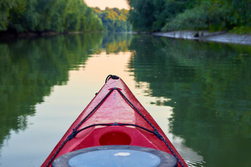 Kayaking near overgrown shore of green thick thickets of trees. Bow of red kayak on Danube river at the morning.
