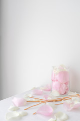 Close up romantic pink rose petals in a glass jar and wicker reed rattan stick on white table top with white wall background with copy space 