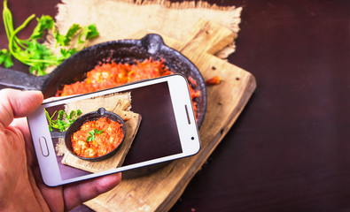 Man takes picture of meatballs in tomato sauce garlic and parsley in an iron frying pan on a dark brown wooden background on smartphone. Copy space