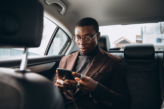 Young smiling african man using smartphone while sitting on backseat in car. Concept of happy business people traveling.