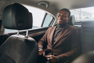 Young smiling african man using smartphone while sitting on backseat in car. Concept of happy...