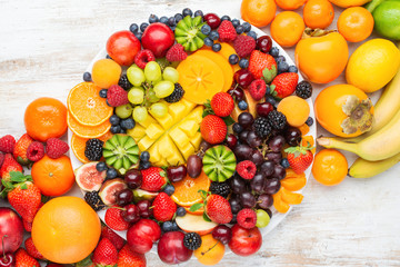 Healthy platter with colorful rainbow fruits, strawberries raspberries oranges plums apples kiwis grapes blueberries mango persimmon, top view, copy space
