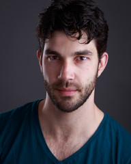 Headshot of handsome caucasian man with stubble and black hair and blue t shirt on grey background