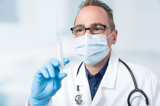 Arzt mit Mundschutz und Stethoskop hält eine aufgezogene Spritze in der Hand