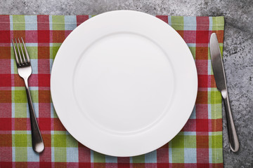 Empty ceramic plate of white color and cutlery on a checkered tablecloth, top view.