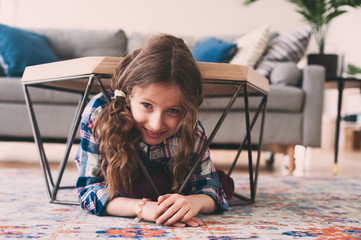 funny naughty kid girl hiding under coffee table at home. Child playing and having fun