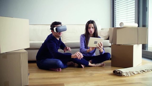 A young couple with tablet and VR goggles sitting on a floor, moving in a new home.
