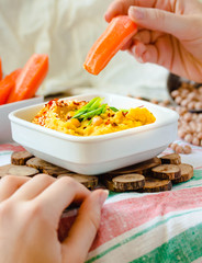 Classic hummus with herbs, olive oil in a ceramic bowl, served with carrot sticks. Vintage background. Traditional Middle Eastern cuisine. Hands of young woman. Eating process.