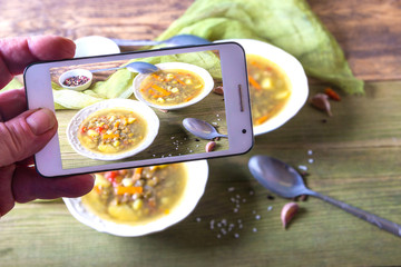 Man takes picture of lentil soup in white plates on wooden background on smartphone. Copy space