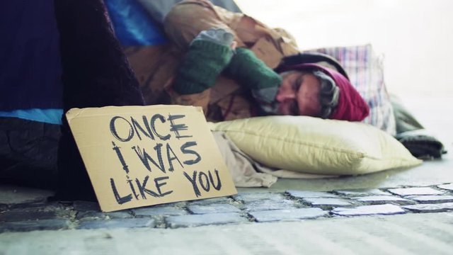 Homeless beggar man lying outdoors in city, cardboard sign near him.