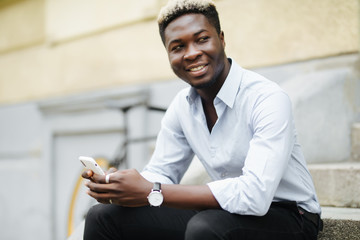 Handsome young Afro American man in casual wear is using smartphone while sitting on stairs outdoors