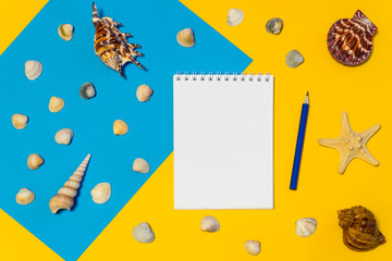 Notebook and pencil with seashells and starfish on blue and yellow paper background. Mock up, flat lay, copy space, top view. Summer vacation concept