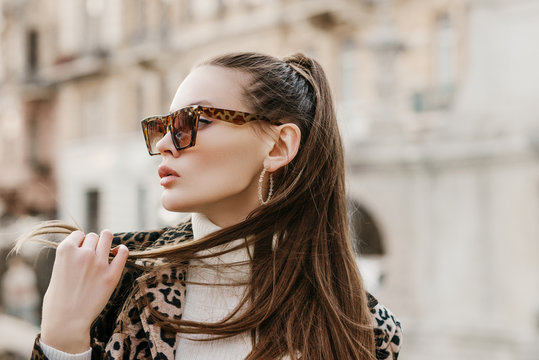 Outdoor close up fashion portrait of young beautiful fashionable woman wearing stylish animal, leopard print sunglasses, hoop earrings, turtleneck, posing in street of european city. Copy, empty space