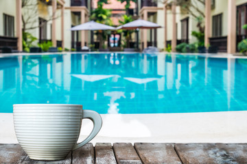 A white cup of coffee on the wooden table by the pool