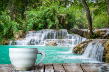 Naklejka premium A white cup of coffee on the wooden table by the warterfall