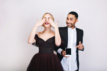 Pretty young woman in luxury evening dress closes eyes with hands, waiting for surprise from handsome guy with ring smiling to camera behind on white background. Valentine s day, proposal, lovers