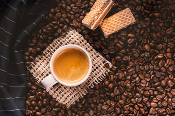 
    Cup of coffee in the middle of coffee beans with biscuits and tablecloth. Grained product. Hot drink. Close up. Harvesting. Natural background. Energy. Fresh espresso with beans on the foam.