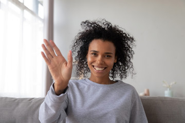 Smiling African American millennial woman sit on couch at home waving to camera, having video call...