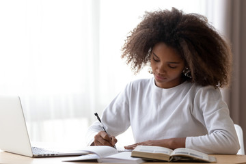 Focused African American teenage girl sit at table with laptop studying with handbooks, serious concentrated black teenager do homework at home, write down in notebook, using computer and textbook - Powered by Adobe