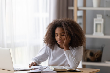 Dreamy African American teenage girl sit at table with laptop and textbooks, distracted from doing...