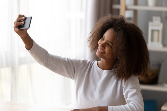 Happy African American teenager hold smartphone, making selfie sitting at table at home, smiling black teen girl using mobile phone posing at camera, taking self-portrait picture on cellphone