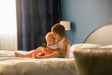 Two sibling children, baby boy and his older brother, sitting on bed in sunny bedroom, playing together