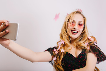 Adorable young girl with blonde long curly hair taking selfie at party, smiling to camera, covered with pink stars confetti. Wearing colorful glasses, black dress. Isolated background.