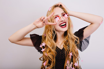 Closeup portrait of cheerful girl with blonde curly hair having great time at party, having fun, celebrating, showing peace to camera. She wearing black dress, pink stylish glasses. Isolated.