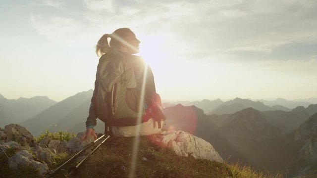 SLOW MOTION, LENS FLARE, PORTRAIT: Young woman sits on a rock and looks at the breathtaking mountains. Bright summer sunbeams shine on the smiling woman resting during a hiking trip in the Alps.