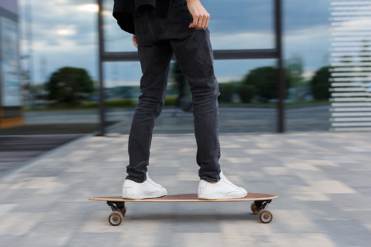 Cropped picture of male in black jeans and white sneakers riding a longboard to the right in urban area, hurry on a date, photo in motion/ side view/ wooden longboard, moving fast, city center.
