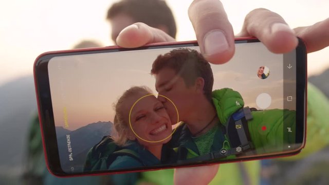 SLOW MOTION, MACRO, DOF: Happy man takes a selfie of him kissing girlfriend on the cheek on a sunny summer evening after reaching the mountaintop in the breathtaking Alps. Hiker couple taking photos.