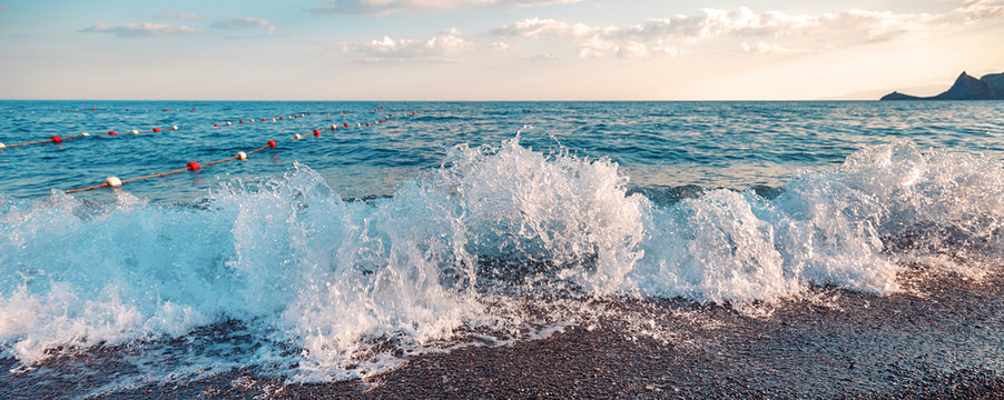 Fototapeta Sunset at rocky beach. Soft evening light reflecting on sea water and small waves with foam, panoramic banner.