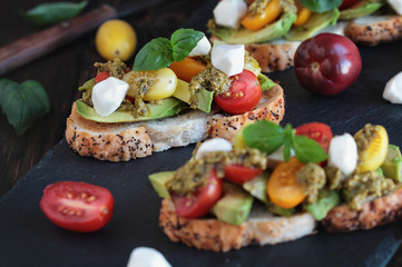 Fototapeta na wymiar Avocado toast sandwich with avocados, pesto, mozzarella cheese, fresh from the garden basil and heirloom tomatoes, over a rustic wooden background. Greek food and healthy vegetarian diet concept.