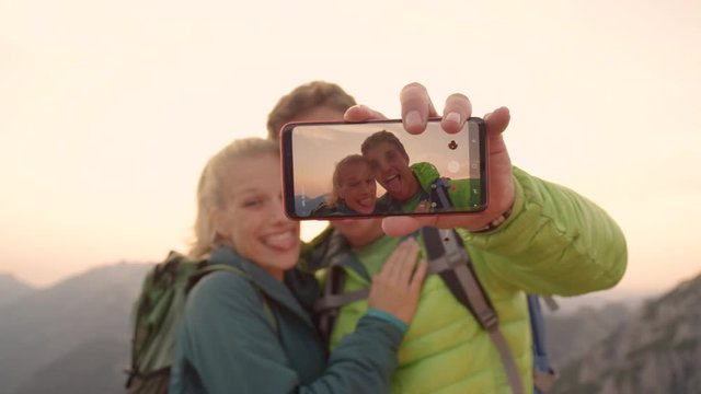 CLOSE UP, DOF: Carefree hiker couple making funny faces while taking a selfie after reaching the mountaintop at sunrise. Happy woman and man taking a photo of themselves during a hiking trip in Alps.