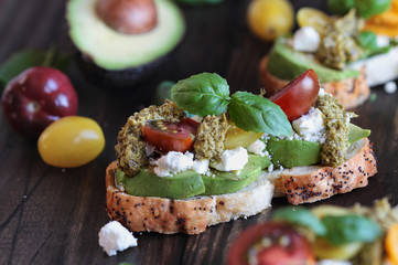 Avocado toast sandwich with avocados, pesto, feta cheese, fresh from the garden basil and heirloom tomatoes, over a rustic wooden background. Greek food and healthy vegetarian diet concept.