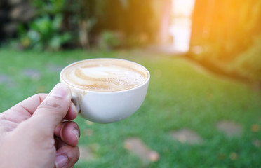 Close-up shot of Hot Latte Fresh Coffee in a white cup in hand with milk foam and smoke, in natural background on the morning with sunrise. Drink concept
