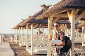 Portrait of a young guy eating a banana with a laptop in his hands standing on a summer canopy of straw. The concept of combining leisure and work in the summer