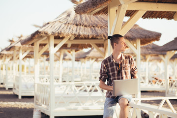 Happy young student guy working with a laptop while sitting under a straw canopy on a warm summer morning. The concept of studying and preparing for summer exams