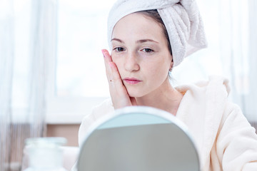 Beautiful happy woman with a towel on her head looking at her skin in a mirror. Timely hygiene and care for the skin